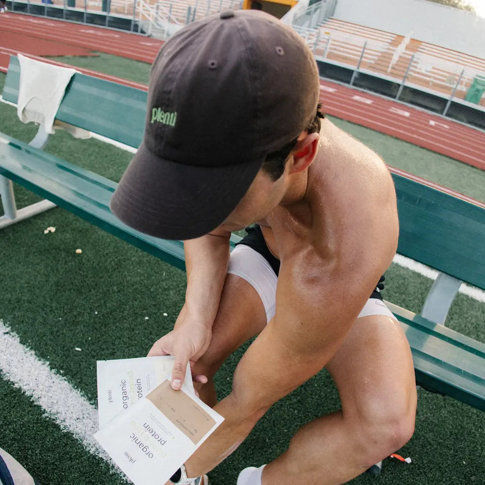 A shirtless man crouching on an athletic field bench wears a dark brown baseball cap with green 'Pleati' embroidery. He is holding two white paper packets of Pleati Organic Protein Powder.