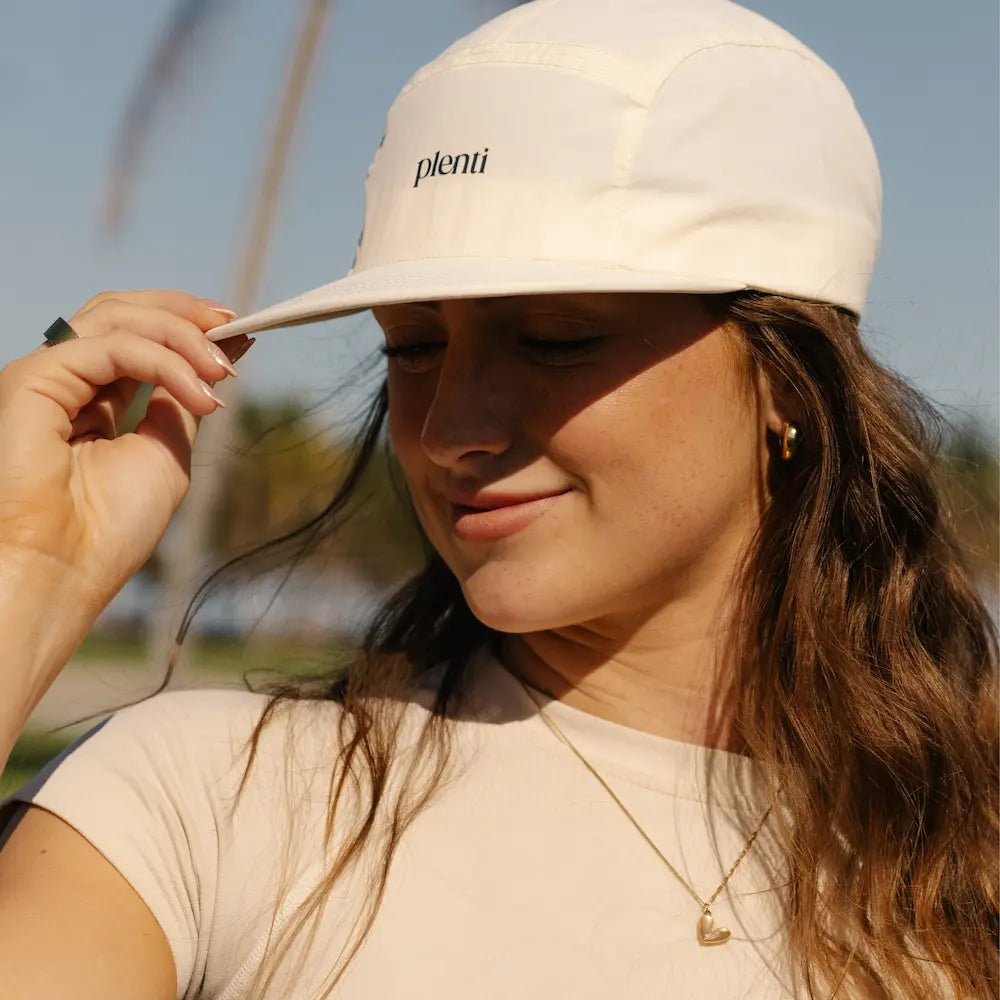 A woman outdoors wearing a cream-colored Plenti cap and a matching cream t-shirt, adjusting the brim of the cap with her hand.