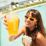 A woman smiling by a swimming pool holds up a clear plastic cup filled with an orange-yellow drink. The cup has the word 'plenti' printed vertically in white.