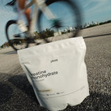 A white stand-up pouch of planti creatine monohydrate sits on asphalt with a blurred cyclist and city skyline in the background.