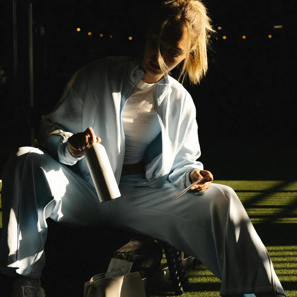 A woman sits outdoors in dramatic low sunlight wearing a light blue oversized zip-up jacket and matching wide-leg pants over a white top. She holds a white bottle, with dark grass and string lights visible in the background.